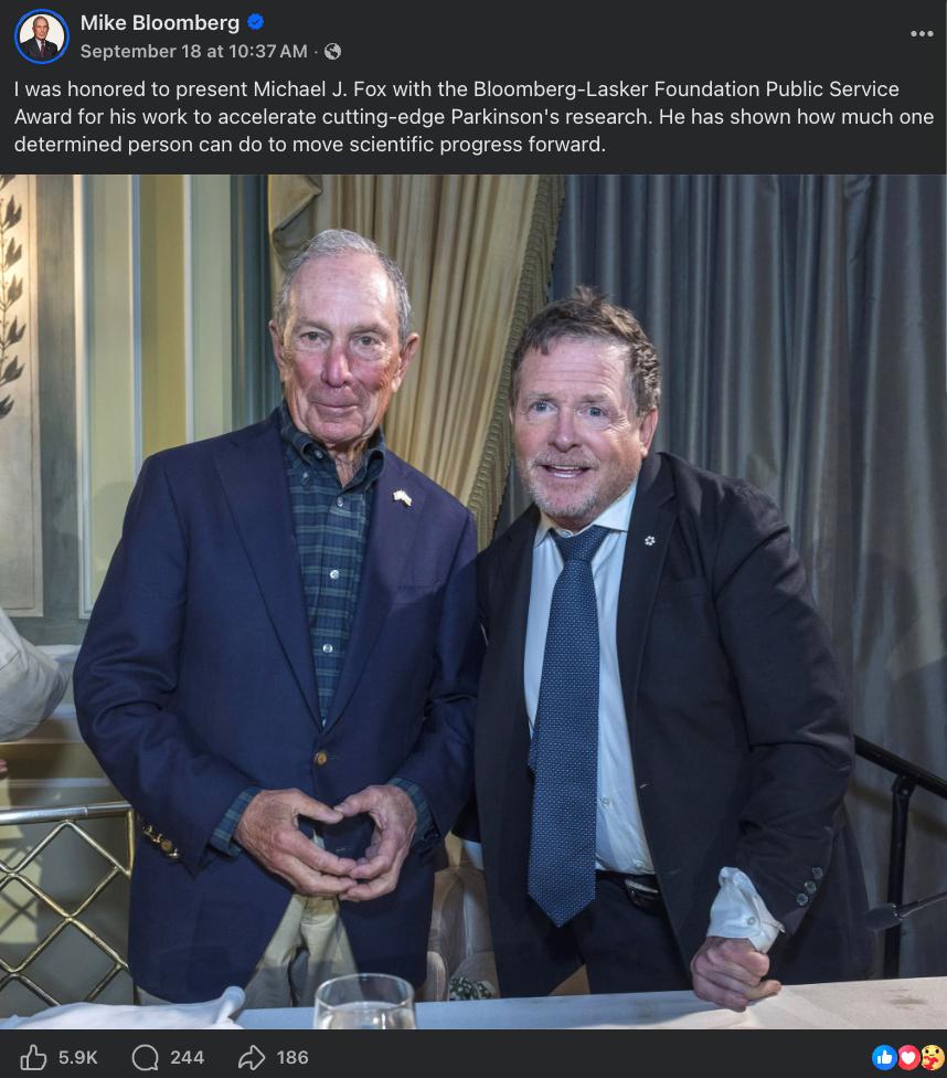 Screenshot of a Mike Bloomberg Facebook post about presenting Michael J. Fox with the Bloomberg-Lasker Foundation Public Service Award, with a photo of Bloomberg in a navy blazer forming a triangle with his hands while standing next to Michael J. Fox.