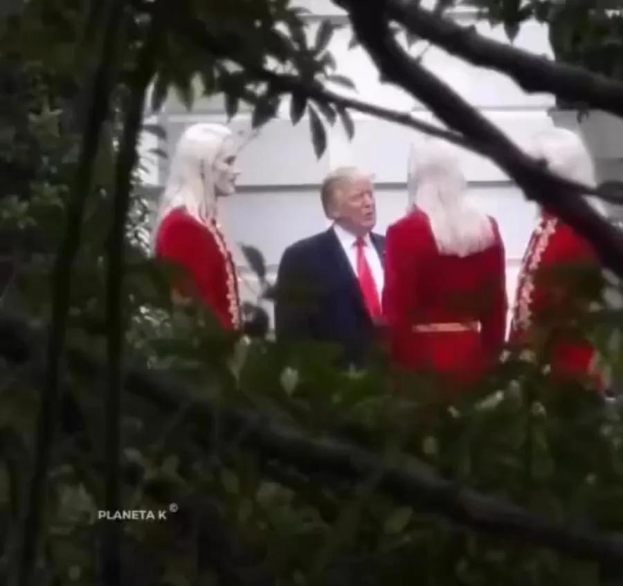 Video still of Donald Trump in a dark suit and red tie standing outdoors between tall figures with long white hair in red embroidered coats, filmed through tree branches near a white building, watermarked Planeta K.